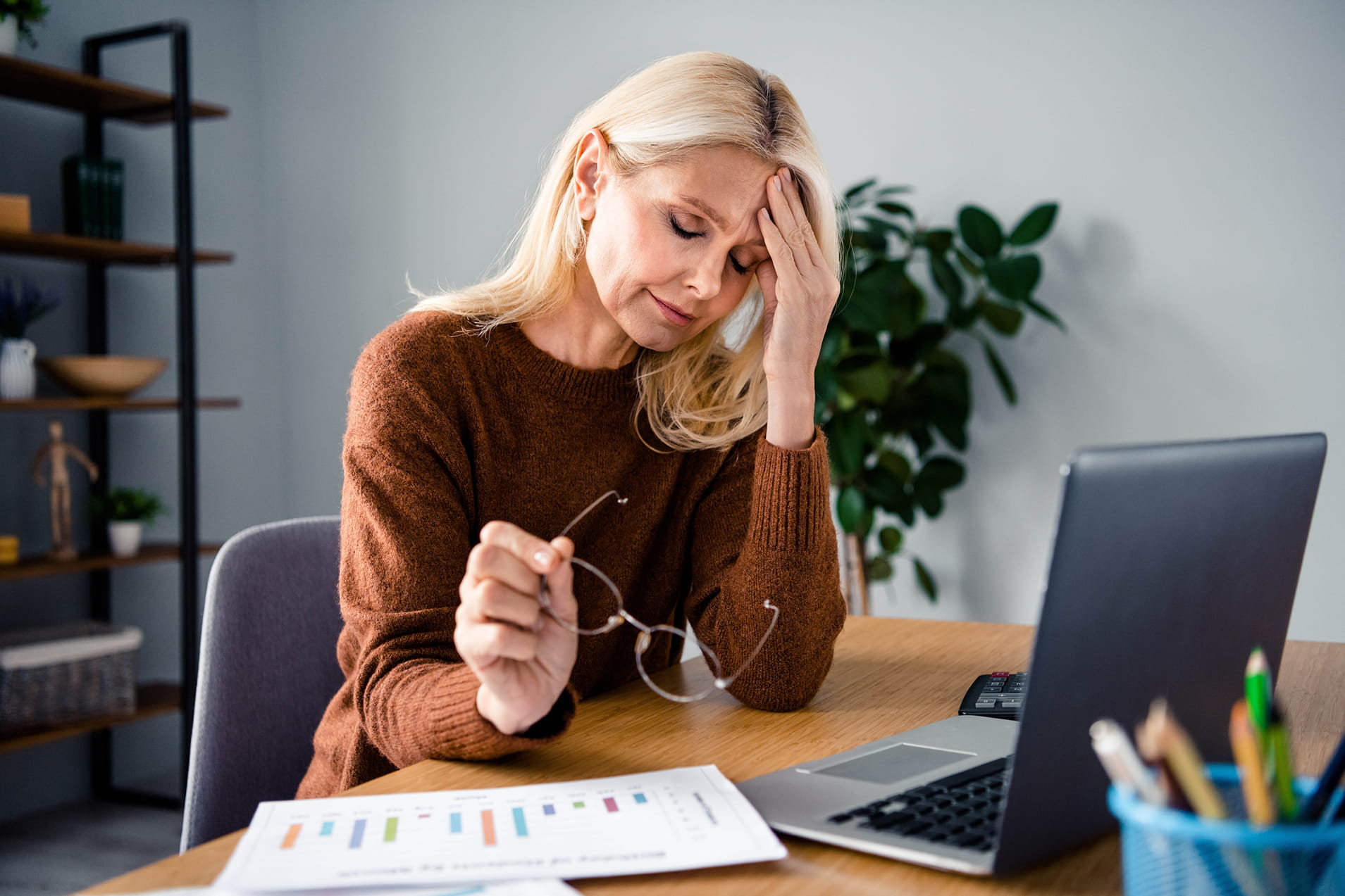 Middle-aged woman looking fatigued and stressed at her desk holding her glasses, representing the brain fog, mood changes and daytime exhaustion caused by menopause-related insomnia — and the need for naturopathic support at the Adelaide Health Clinic
